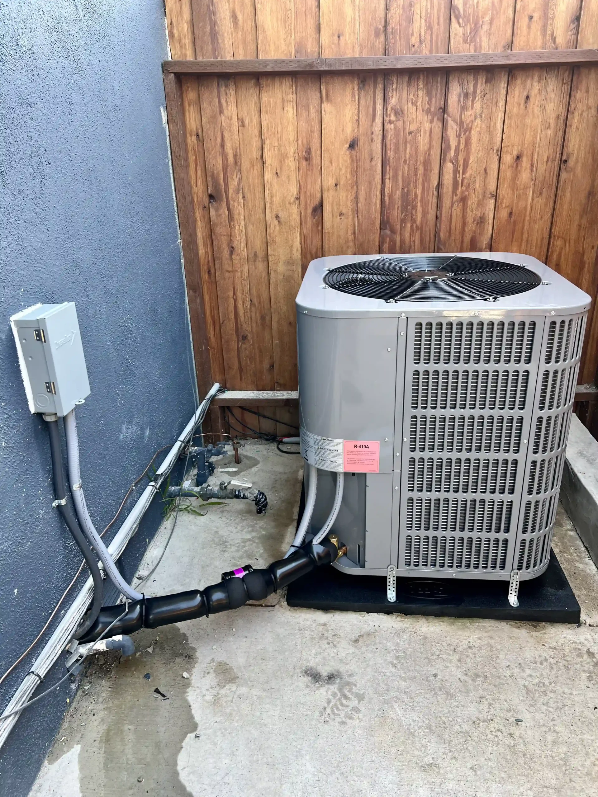 An outdoor central air conditioning unit positioned between a fence and a blue stucco wall in Riverside.