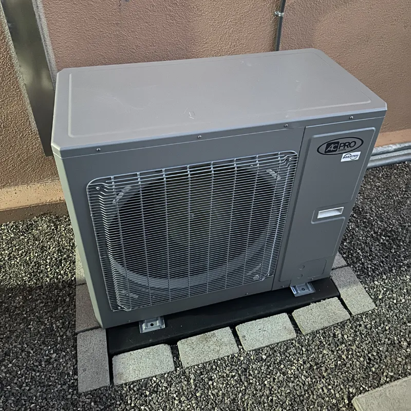 A grey AC Pro inverter heat pump unit sitting outside a stucco wall on a custom paver and gravel base.