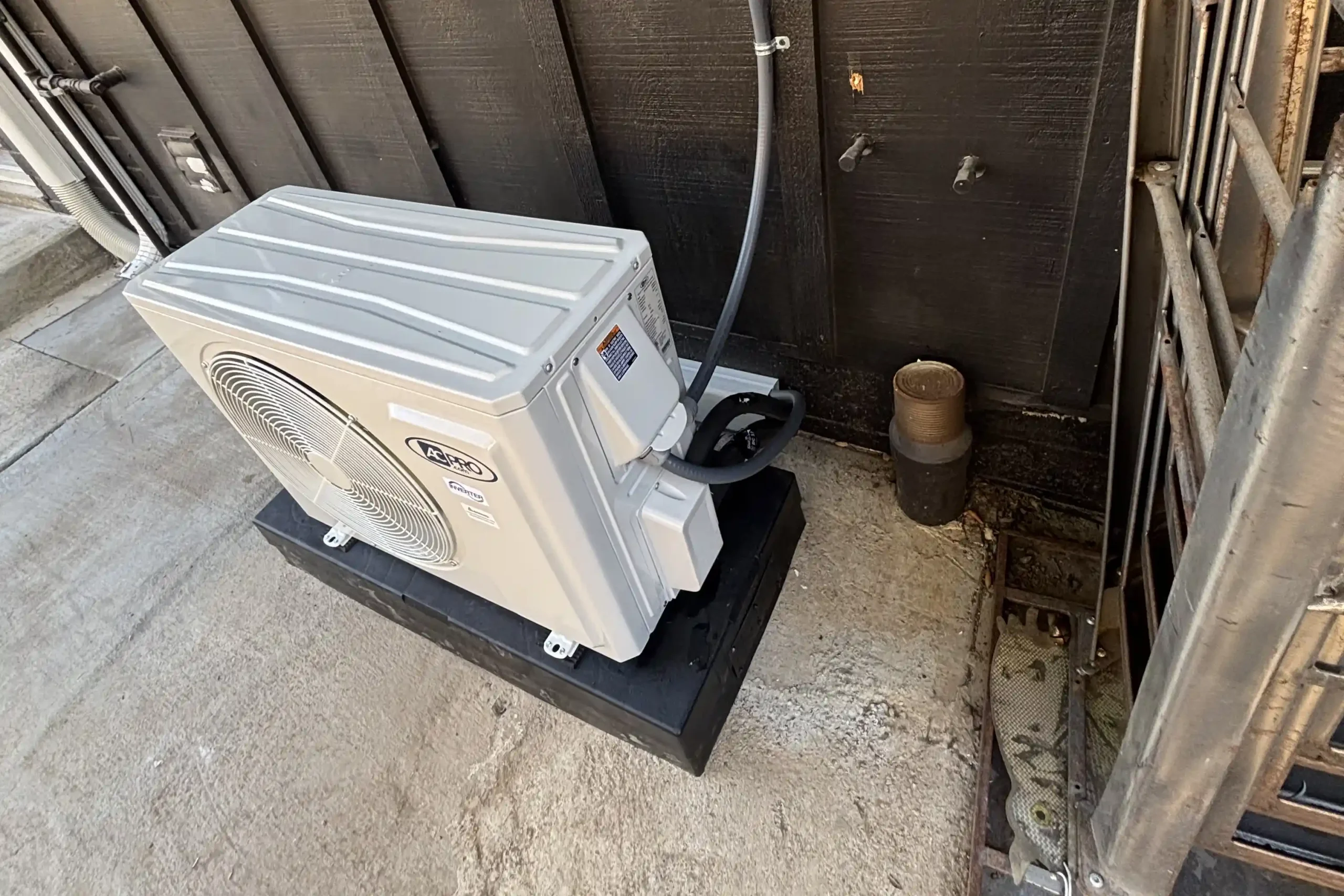 A white AC Pro ductless mini split outdoor unit installed on a black pad against a dark wooden wall in Huntington Beach.