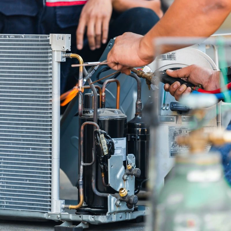A technician using a torch to weld and secure copper line connections on an air conditioning unit frame.