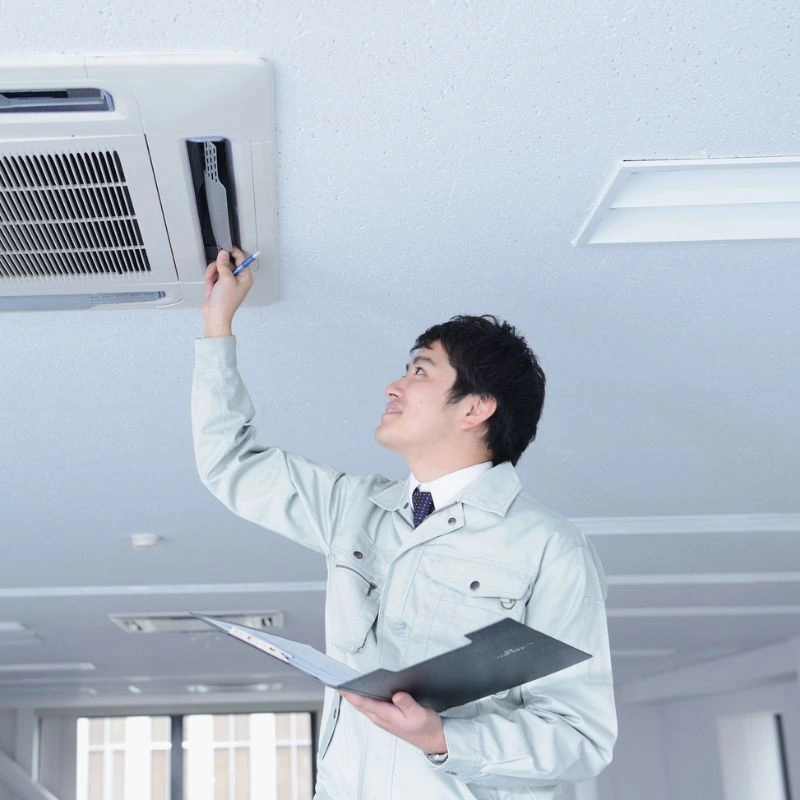 An HVAC professional holding a clipboard while performing a detailed cooling system inspection on a ceiling cassette unit.