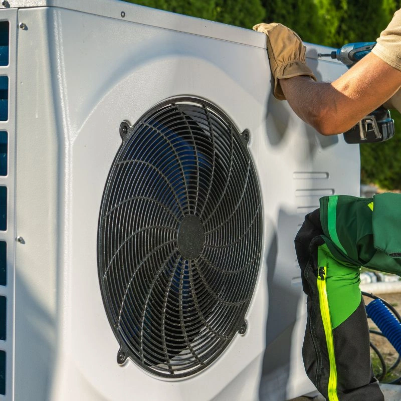 A close-up of a technician using a cordless drill on the side frame panel of a white outdoor heat pump unit.