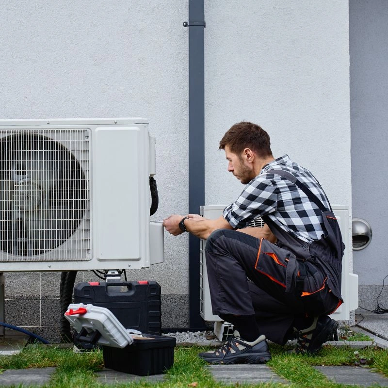 An installer checking exterior conduit connections on a wall-side residential air compressor unit during a service call.